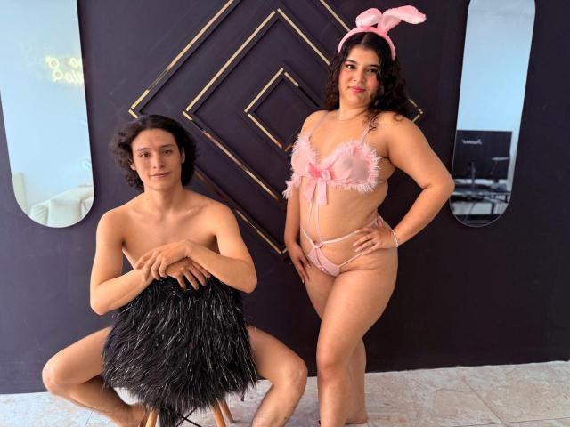 LizAndDante with curly brown hair, wearing pink frilled lingerie with bunny ears, stands next to Dante, who is shirtless, holding black pom-poms, in a room with dark walls.