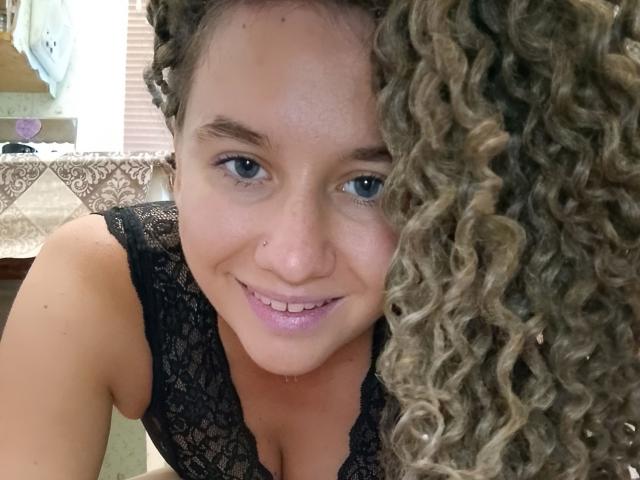 MarlanViviane has curly brown hair, blue eyes, a nose piercing, and is smiling at the camera with pink lipstick. She's wearing a black lace top, showing her tits.