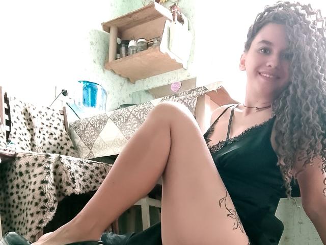MarlanViviane with curly brown hair, smiling, wearing a black satin camisole, sitting with legs crossed, showing a tattoo on her right thigh, in a bright room.