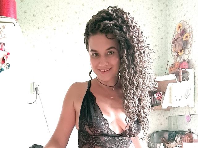 MarlanViviane with curly gray and brown hair, wearing a black lace bra, smiling, showing her tits, in a bright room with a white fridge and shelf on the right.