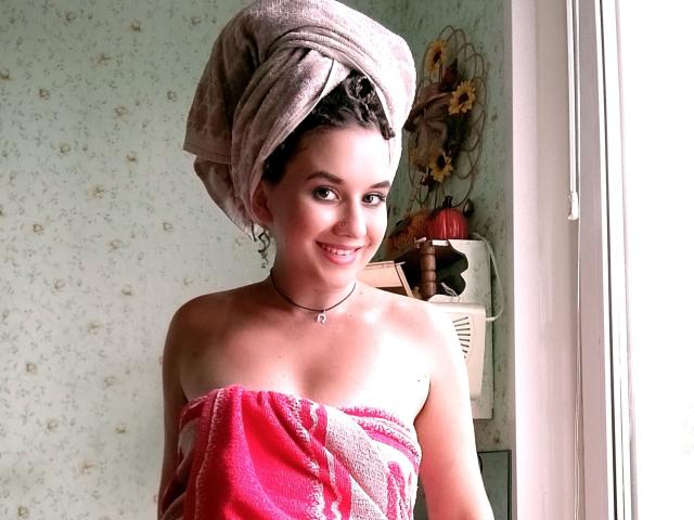 MarlanViviane with a towel wrapped around her boobs, pink and white towel, brown hair in a towel, smiling, standing by a window, necklace, floral wallpaper, lamp in background.