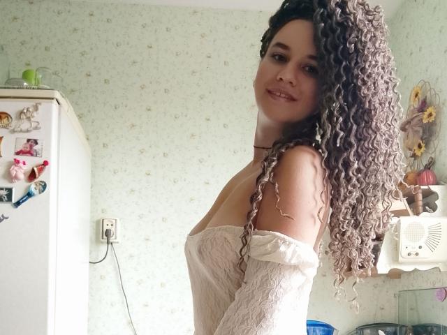 MarlanViviane with curly, grayish-brown hair, wearing a white, off-shoulder lace top, smiling at the camera, showing her tits and a cute face, in a kitchen.