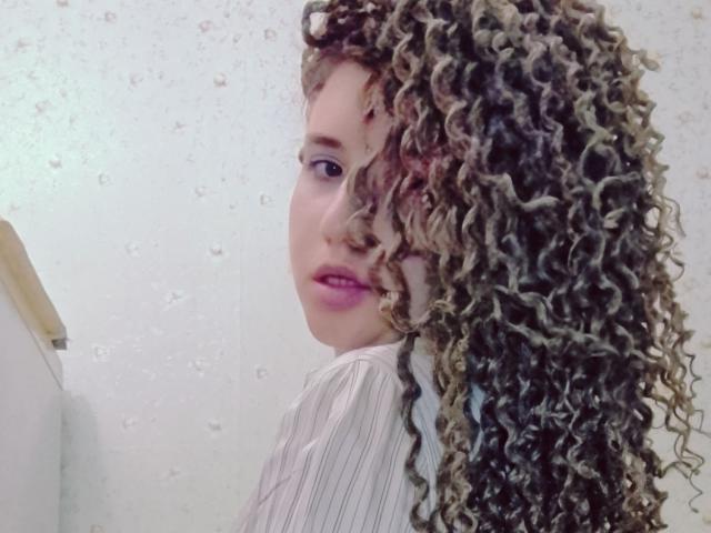 MarlanViviane with curly brown and blonde hair, pink lipstick, looking over shoulder, wearing a striped shirt, white wall with water droplets in background.