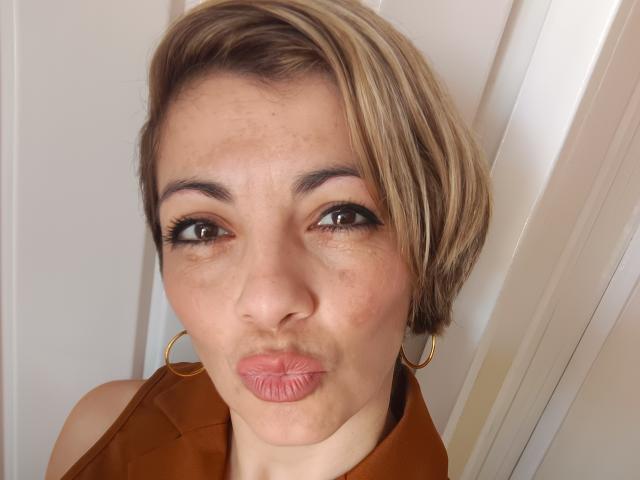 ChanelJolie with short blonde hair, brown eyes, and gold hoop earrings, making a pouty kissy face, wearing a brown top, showing freckles on her fair skin.
