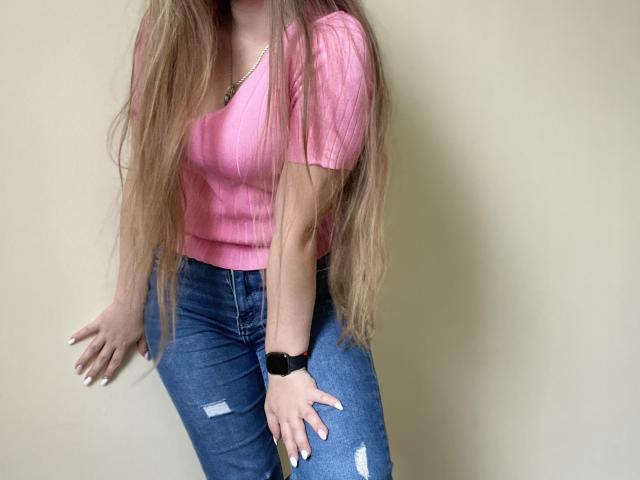 Doris met lang, rechtbruin haar, een roze geribbelde top en blauwe, versleten jeans, draagt een zwarte polshorloge en staat tegen een effen muur..