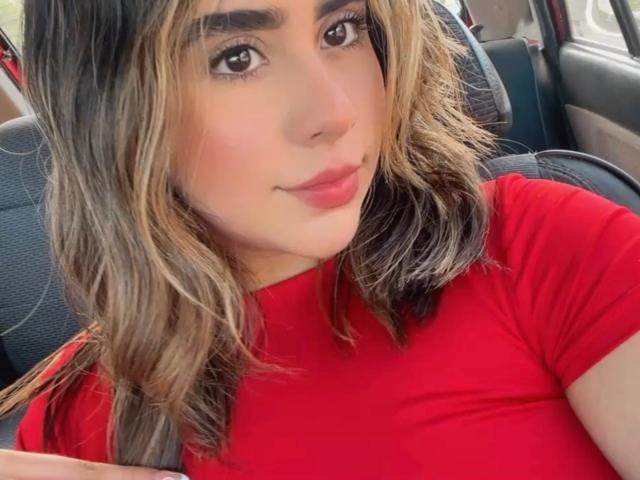 OlivaStellas with wavy blonde and brown hair, wearing a red turtleneck, has pouty red lips and brown eyes, sitting in a car, looking at the camera.