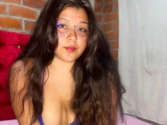 CurvyHotElaine with long black hair, wearing round glasses and a blue bra, showing her big tits and pink lips, sitting against a red and white wall.