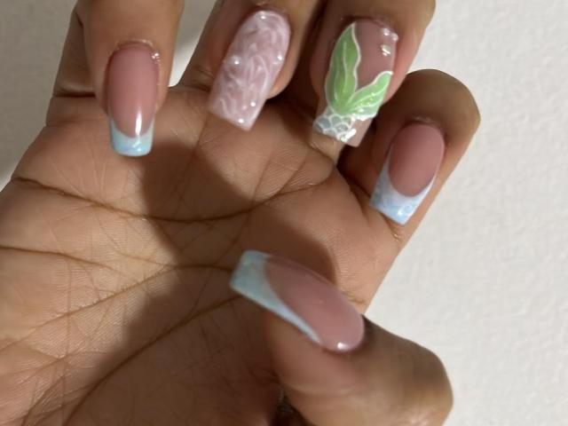 SiennaTaylor's nails are glossy with pink and blue French tips, one's pink with glitter, another's green leaf design, and the rest are plain pink, showing off her manicured hand.