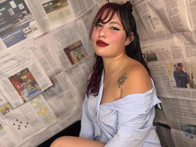 DominicTheOne with dark and red-dyed hair, off-shoulder light blue shirt, red lipstick, tattoo on left shoulder, sitting against newspaper-covered wall, looking at camera with a smile.
