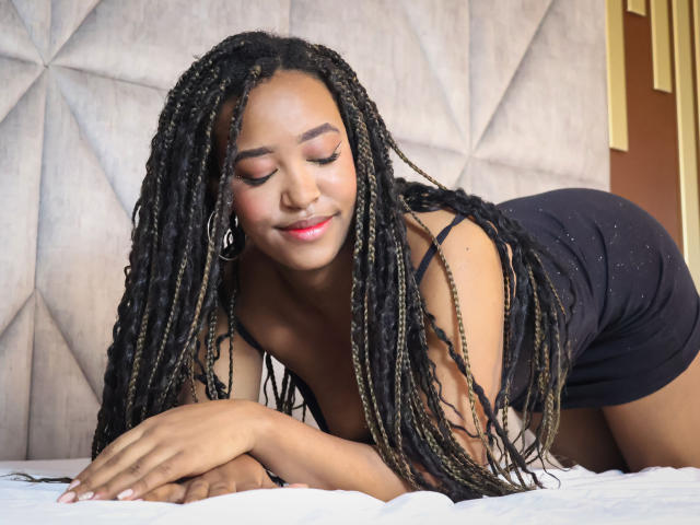 NaomiReyes with long black and blonde braids, wearing a black tank top and shorts, on all fours on a bed, smiling with eyes closed, showing her ass and breasts.