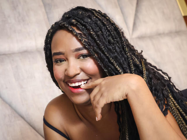 NaomiReyes with dark brown skin, smiling, showing white teeth, red lipstick, black braided hair, finger resting on chin, background is blurred, close-up, head and shoulders.