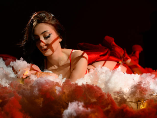 AnyaLobb with red ribbons on her tits, lying in fluffy white and red smoke, eyes closed, dark hair, red lingerie, and red ribbon on her ass, in a dark setting.
