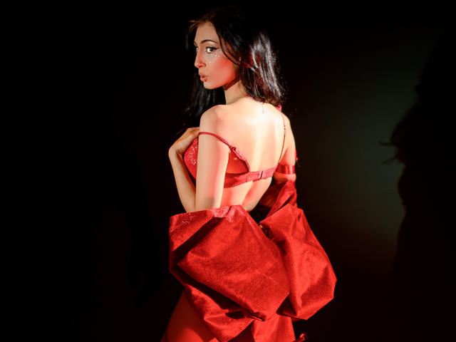 AnyaLobb with dark hair, red lace bra, and red satin dress slipping off, showing her bare back and tits, looking over her shoulder, in a dark, lit-up photo.
