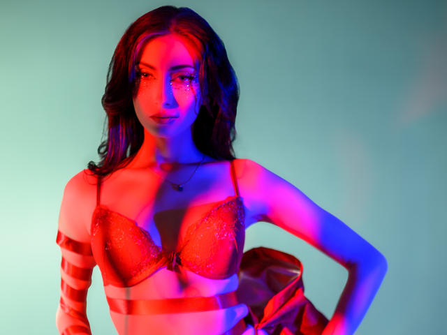 AnyaLobb with red bra, glittery, wearing a red sheer top, hands on hip, wavy brown hair, blue and red light, looking confident, slight smile, pink nipples visible.