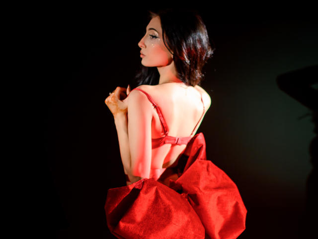 AnyaLobb with red bra and red dress, showing her back and bare ass, holding her dress, dark hair, black background, soft light, sexy, elegant pose.