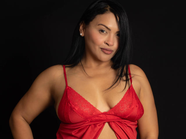 EstrellaSmmith, con senos de tamaño medio, lleva un sujetador de encaje rojo y mira a la cámara con una ligera sonrisa. Tiene el pelo negro y el fondo es oscuro. Lleva una blusa de satén rojo..