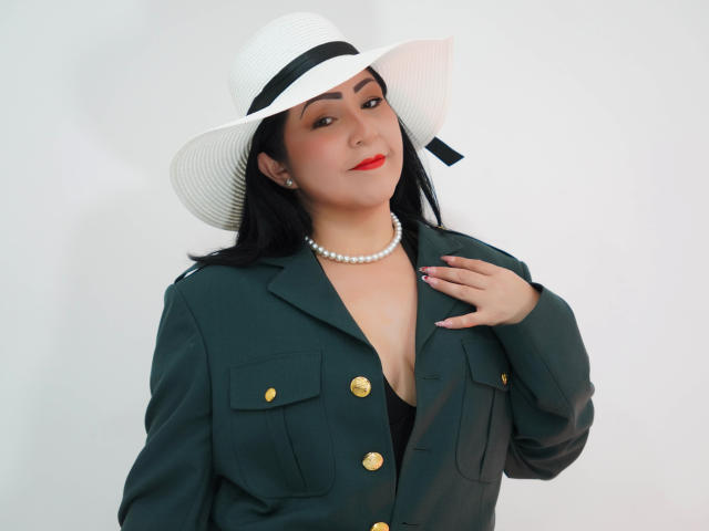 ArabelaFoxy, com cabelo escuro, batom vermelho, colar de pérolas, jaqueta verde, vestido preto, chapéu branco, mão no peito, mostrando o decote, olhando para a câmera, fundo branco..