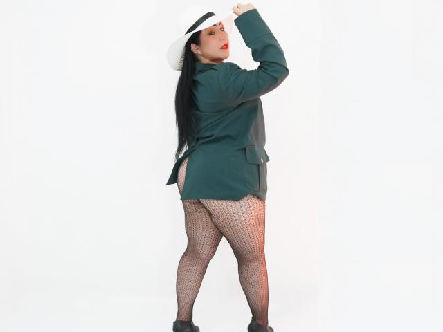 ArabelaFoxy staat met haar rug naar de camera, draagt een groene jas, een witte hoed, zwart haar, rode lippenstift, gestreepte leggings, zwarte hakken, en haar achterkant is zichtbaar. Ze heeft lange benen en een rond figuur..