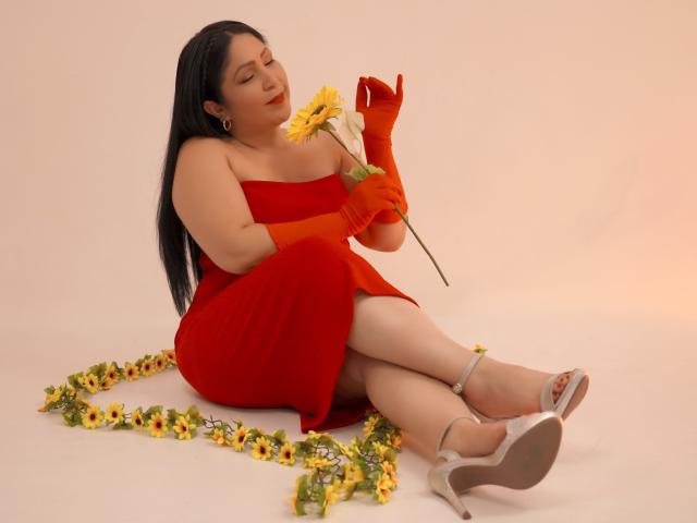 ArabelaFoxy met lang zwart haar, een rode jurk, oranje handschoenen, die een zonnebloem vasthoudt, zittend op een witte vloer, omringd door gele bloemen, met witte hakken, ogen gesloten, lachend..