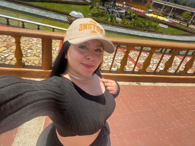 EmaJames has big boobs, wearing a black ribbed crop top, beige cap, and silver choker, standing on a red brick balcony, smiling, with long black hair.