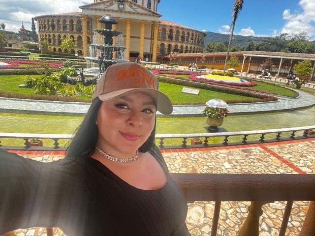 EmaJames with large tits, wearing a brown cap and black top, smiling in a sunny garden with a fountain and ornate building. She has green eyes and a necklace.