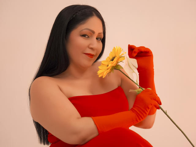 ArabelaFoxy, avec de longs cheveux noirs, une robe rouge sans bretelles et des gants orange, tenant un tournesol jaune près de son nez, souriante, avec du rouge à lèvres rouge, une peau claire, et ayant une allure à la fois mignonne et séduisante..