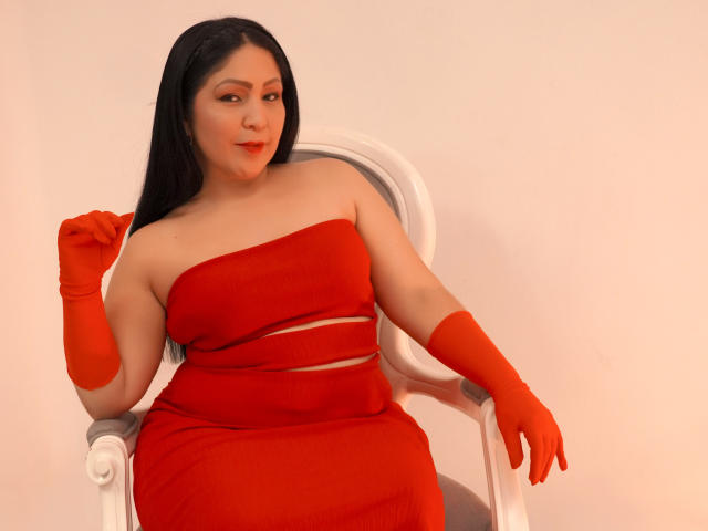 ArabelaFoxy mit langem, schwarzem Haar, die ein rotes, trägerloses Kleid und passende rote Handschuhe trägt, sitzt auf einem weißen Stuhl, leicht lächelnd, die rechte Hand ist erhoben, die linke Hand liegt auf dem Stuhl..
