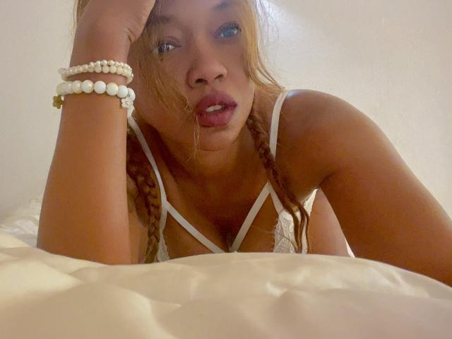 LaPicante with braided red hair, white lace bra, and beaded bracelets, lying on a bed, looking up with parted lips, soft light, tan skin, and blue eyes.