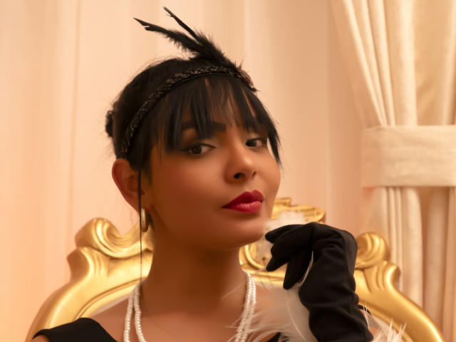 JessicaFiore, met donker haar, een zwarte hoofdband, een zwarte veer, rode lippenstift, zwarte handschoenen, een parelketting en een witte veer, zittend op een gouden stoel, met een zelfverzekerde en aantrekkelijke uitstraling..