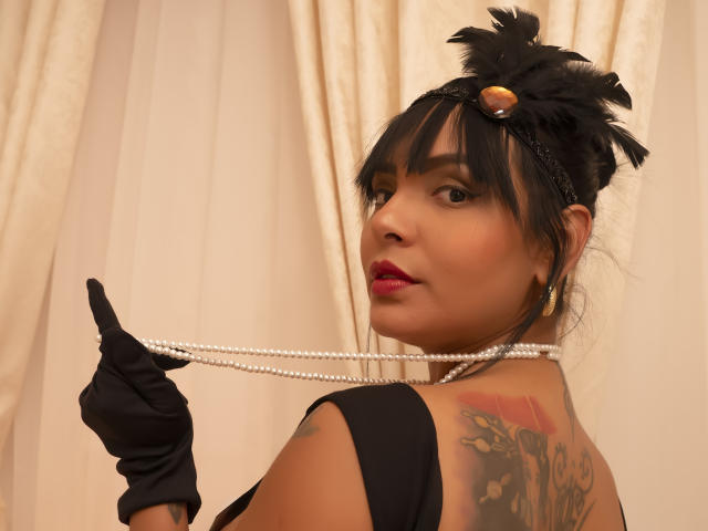 JessicaFiore, avec des plumes noires dans ses cheveux, tenant un collier de perles et portant des gants noirs, montrant un tatouage sur son dos, avec du rouge à lèvres, et regardant par-dessus son épaule..