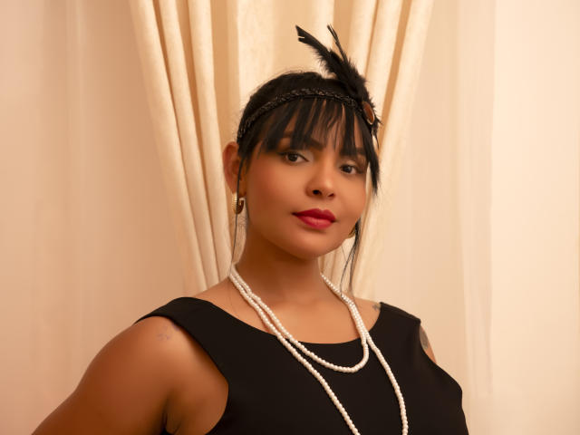 Jessica Fiore, com cabelo escuro, adorno de penas pretas na cabeça, batom vermelho, vestido preto sem mangas, colar de pérolas brancas, brincos pequenos, em frente a cortinas bege, olhando para a câmera..