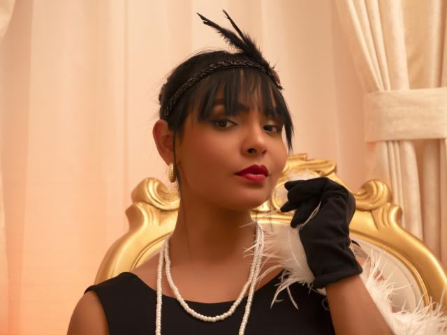JessicaFiore, con capelli scuri, fascia nera per capelli, vestito nero, collana di perle, guanti neri, piuma bianca, rossetto rosso, poggiatesta dorato, che guarda leggermente di lato, tenendo la piuma vicino al viso..