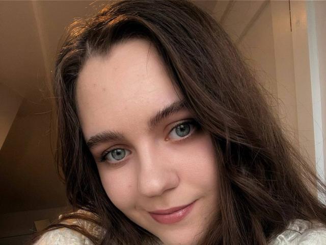 LidiYanna tem olhos azuis, cabelo castanho ondulado e um sorriso discreto. Seu rosto adorável tem uma pequena espinha na bochecha direita. Ela está vestindo uma blusa branca..