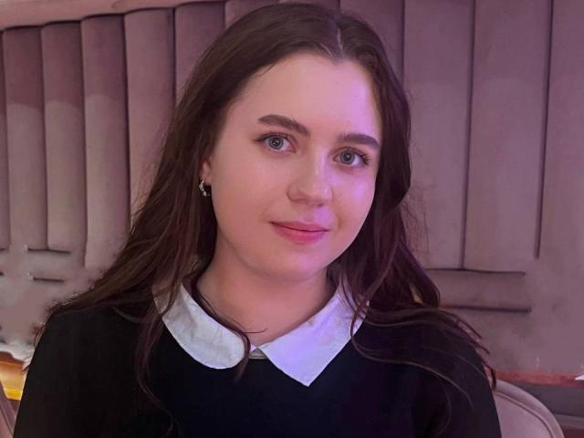 LidiYanna has long brown hair, blue eyes, and a slight smile. She's wearing a black top with a white collar. Her tits are small, and she's looking directly at the camera.
