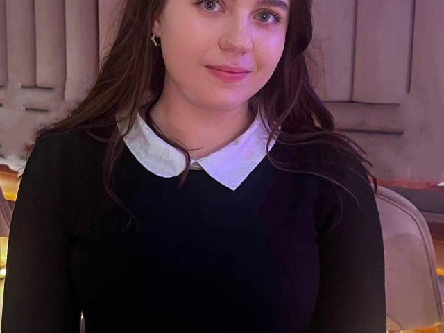 LidiYanna, avec de longs cheveux bruns, des yeux bleus et des lèvres roses, portant une robe noire avec un col blanc, souriant doucement. Les seins ne sont pas visibles, l'accent est mis sur le haut du corps..
