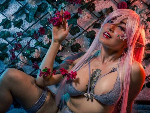 MeiJaisen with pink hair, elf ears, tattoos, wearing lacy grey bra, holding pink flowers, seductive expression, blue and pink lighting, showing tits, sitting with leg up.