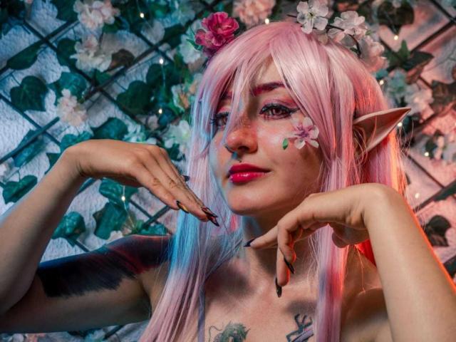 MeiJaisen with pink hair, elf ears, and flower headpiece, showing off nails and tattoos, has pink lipstick and floral makeup, posing with hands up, showing off her tits.