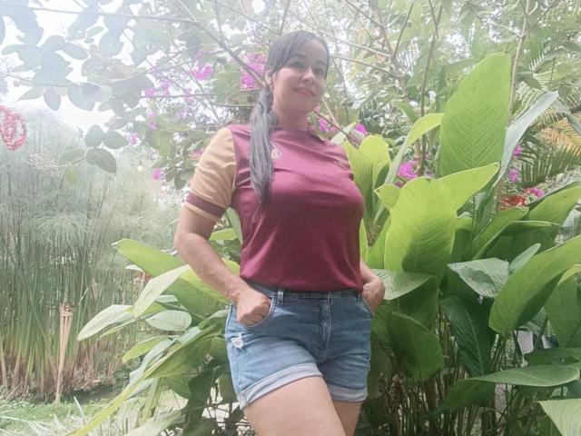 TamaraFoxy stands in a garden, wearing a maroon and tan shirt, blue denim shorts, hands in pockets, green plants, pink flowers, and long black hair, smiling.