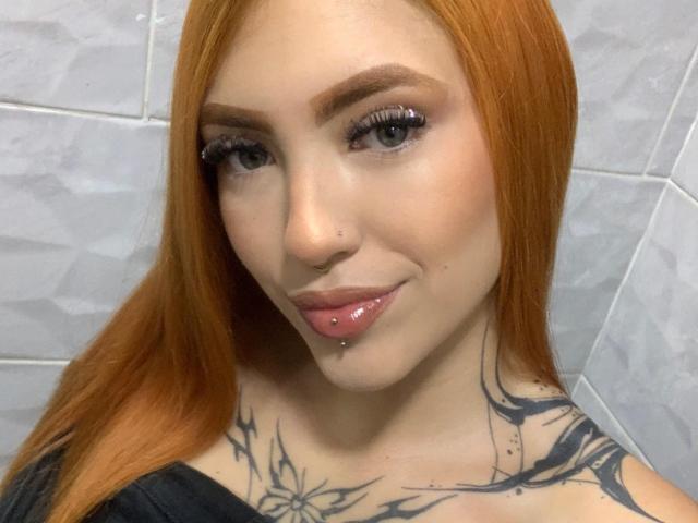 AntonellaBrith has red hair, a nose ring, a lip piercing, and a tattoo with floral designs on her chest. She's smiling with glossy pink lips and long eyelashes.