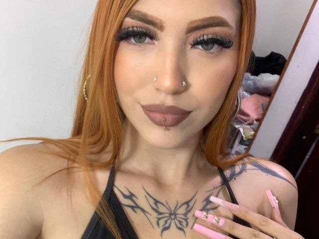 AntonellaBrith has orange hair, green eyes, nose piercings, lip piercings, butterfly tattoo on chest, pink nails, and hoop earrings. Boobs visible in black top. Smiling.