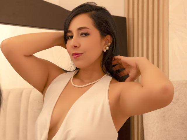 KattyRivera with medium tits, wearing a deep white dress, pearl necklace, and earrings, has black hair, pink lipstick, and a confident smile, arms raised, in a warm-lit room.