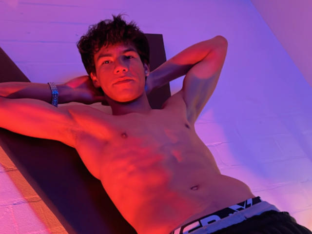 NewmanFire is shirtless with arms behind head, wearing black pants with white waistband, lying on dark surface, red and purple lights on his skin, brown curly hair, bracelet on wrist.