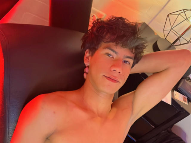 NewmanFire, shirtless, with curly brown hair, blue eyes, and pink ear gauges, lying on a black chair, arm behind head, red and orange light, slight smile, muscular, relaxed.