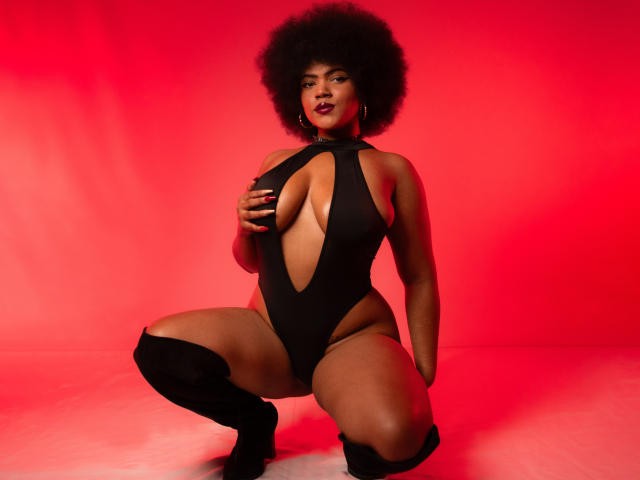 Alice Jelanni, con un afro grande, vestida con un traje de baño negro con cortes, agachada, con una mano en los senos, botas negras hasta la rodilla, fondo rojo, expresión segura, sexy, piel oscura, labios carnosos..