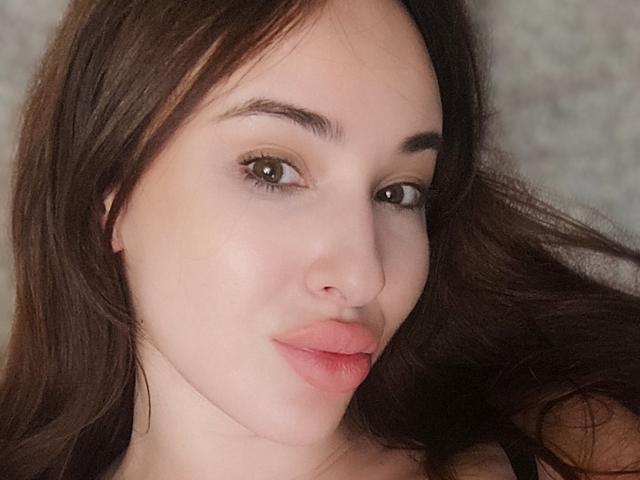WillaAdriana's face, showing light skin, brown eyes, and full pink lips, with long brown hair, and subtle makeup, looking slightly to the right with a soft smile.
