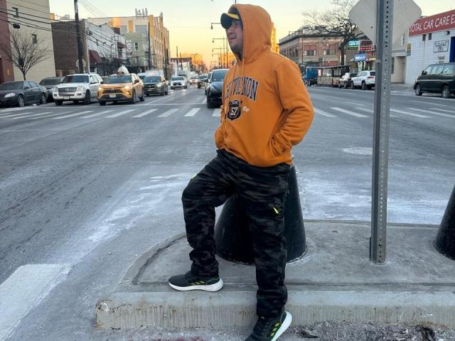 ElianGoldS, wearing an orange Michigan hoodie, black camo pants, and black sneakers, sits on a black dumpster, hands in pockets, with a street and cars in the background.