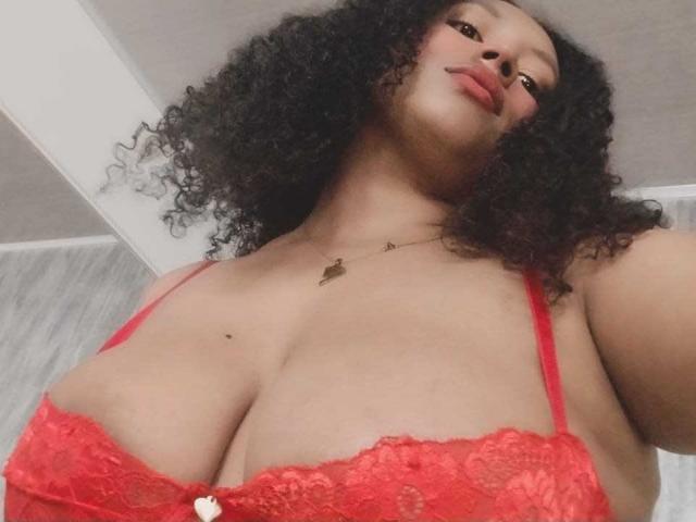 KarinaLovey with curly black hair, red lace bra, large tits, and a small mole on her chest, wearing a gold necklace, looking down at the camera with red lipstick.