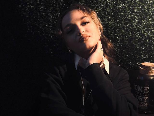 AfinaCrazy with pouty lips, black hoodie, and white shirt, holding her chin, has a tattoo on her wrist, and is next to a jar, in dim light.