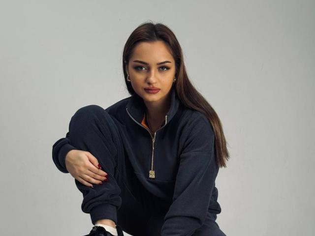AfinaCrazy with long brown hair, dark eyes, and red lipstick, squatting, wearing a dark zip-up hoodie, black pants, and white socks, with a gold necklace.