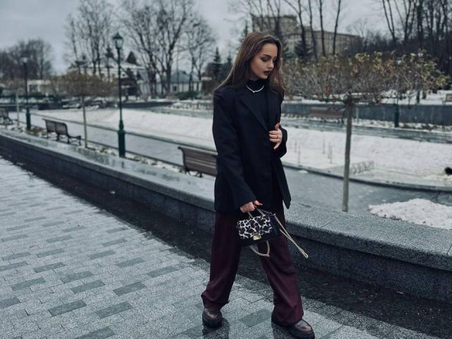 AfinaCrazy stands in a snowy park, wearing a black blazer, maroon pants, brown shoes, and a leopard print clutch, with long brown hair and a serious expression.